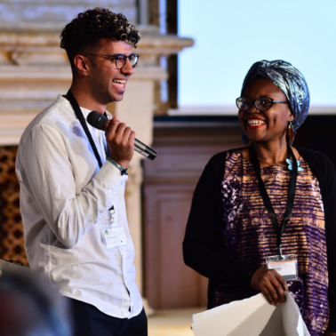 Young BIPOC man with glasses stands with microphone next to BIPOC woman with blue headwrap