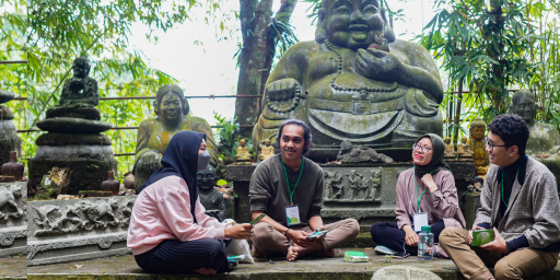 Four Indonesian youth sit in front of a stone statue surrounded by lush greenery, talking