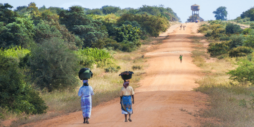 Two African women women with baskets on their heads walk up a dusty road towards a water tower in the distance